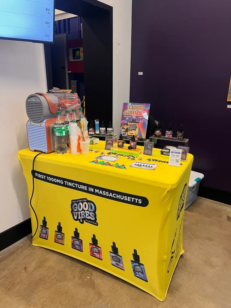 Bright yellow Good Vibes pop-up table with tincture bottles and a frozen-drink station at Mayflower – Allston.