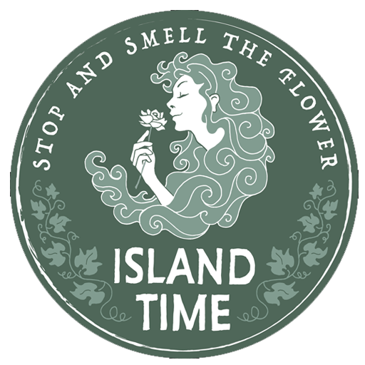 Circular green badge featuring a white line drawing of a woman smelling a flower, with decorative vines and the text 'ISLAND TIME' at the bottom and 'SMELL THE FLOWER' around the edge.